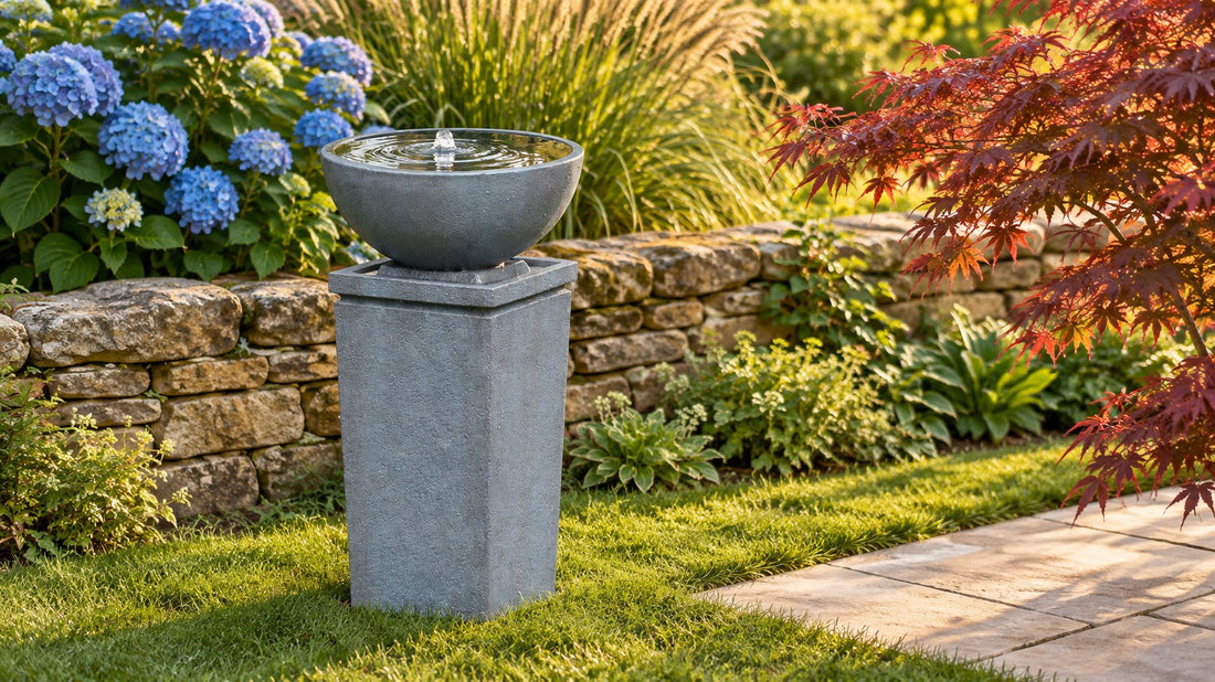 Wide grey bowl on a square plinth holding an almost still surface, hydrangeas and a dry stone wall behind