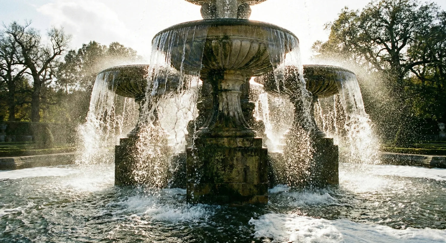 A large, multi-tiered classical stone fountain with heavy water cascading, creating significant splash, mist, and foam, backlit by dramatic sunlight.