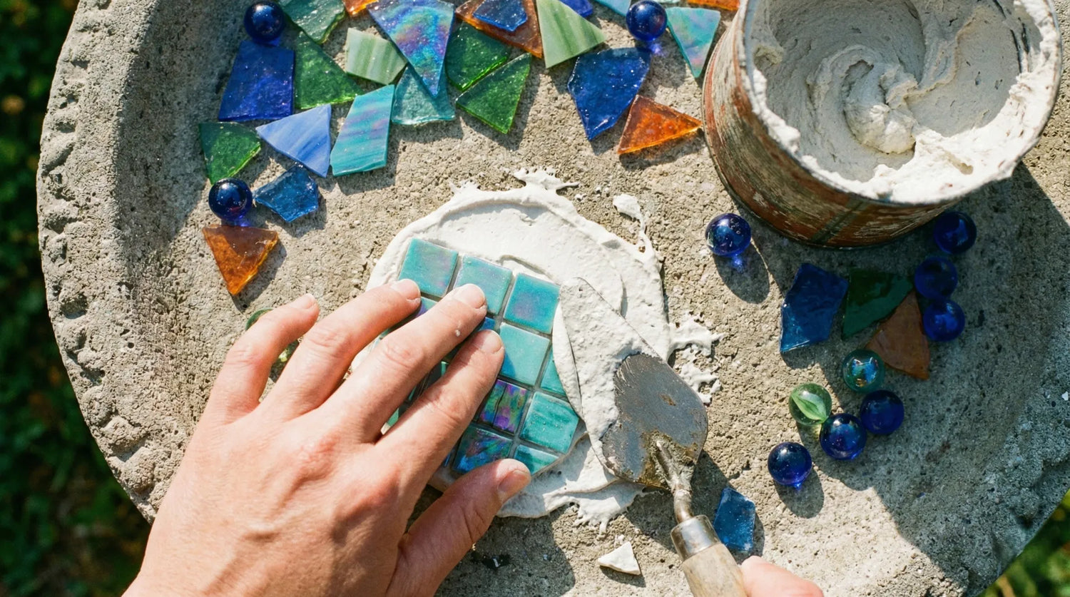 DIY process of making a mosaic birdbath showing a hand placing turquoise glass tiles on a concrete basin with mortar and colorful glass shards.