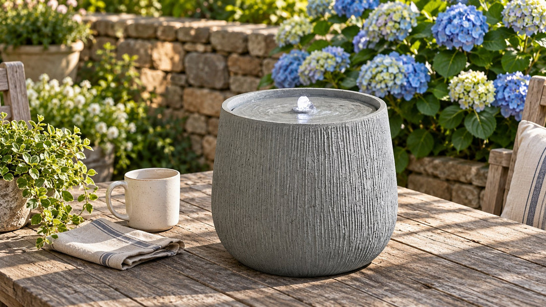 Small ribbed drum fountain on a weathered wooden table with a mug beside it for scale, hydrangeas behind