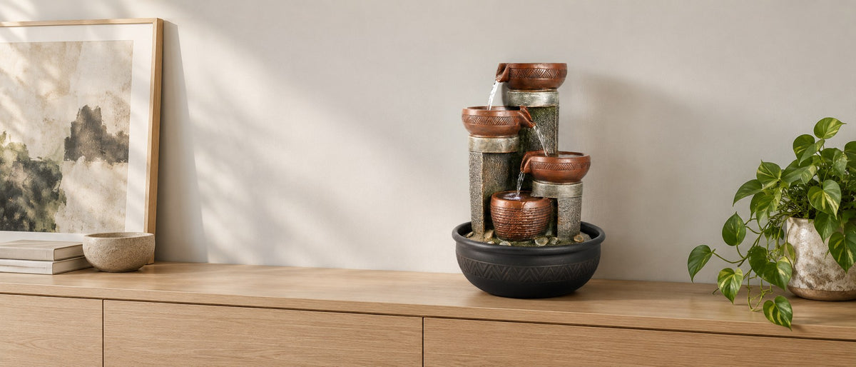Carved terracotta bowl tabletop fountain on a pale oak sideboard with framed art and a trailing plant