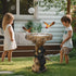 Children watching wild birds fly around a black bear tree stump bird bath in a garden.