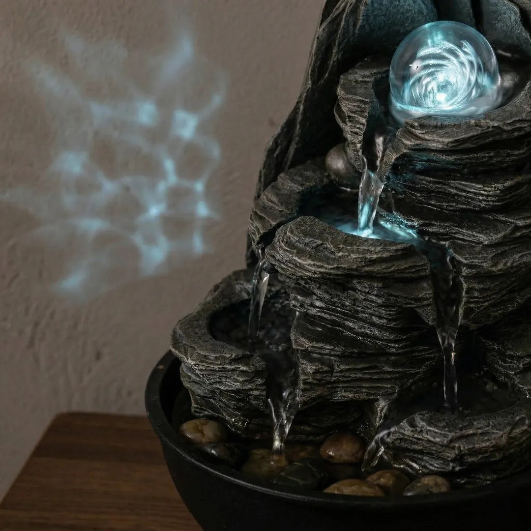 Close-up of a tabletop fountain at night, illuminated by warm LED lighting and casting gentle reflections on a wall.