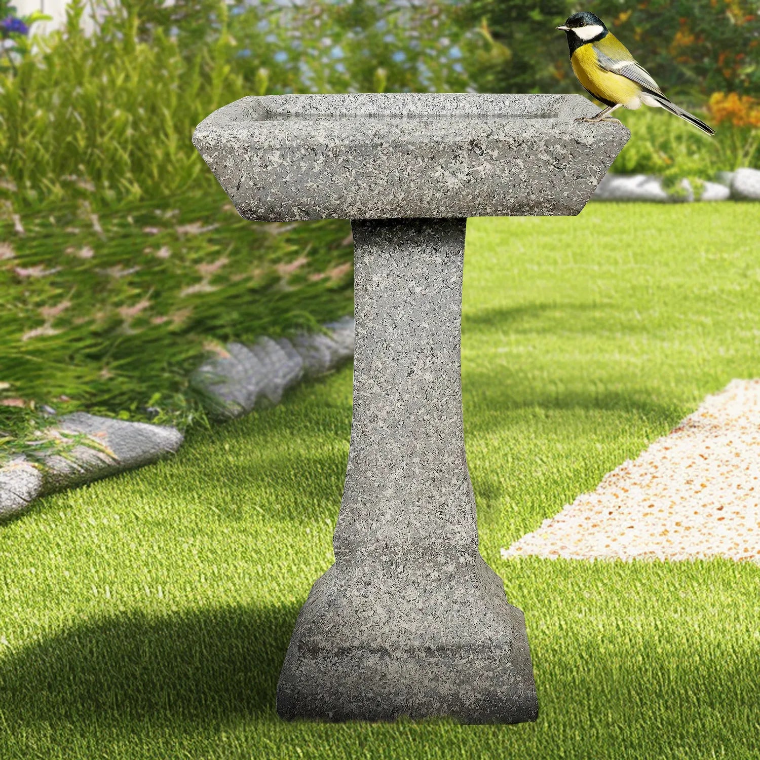 Contemporary faux stone pedestal birdbath placed on a manicured lawn next to a garden path.