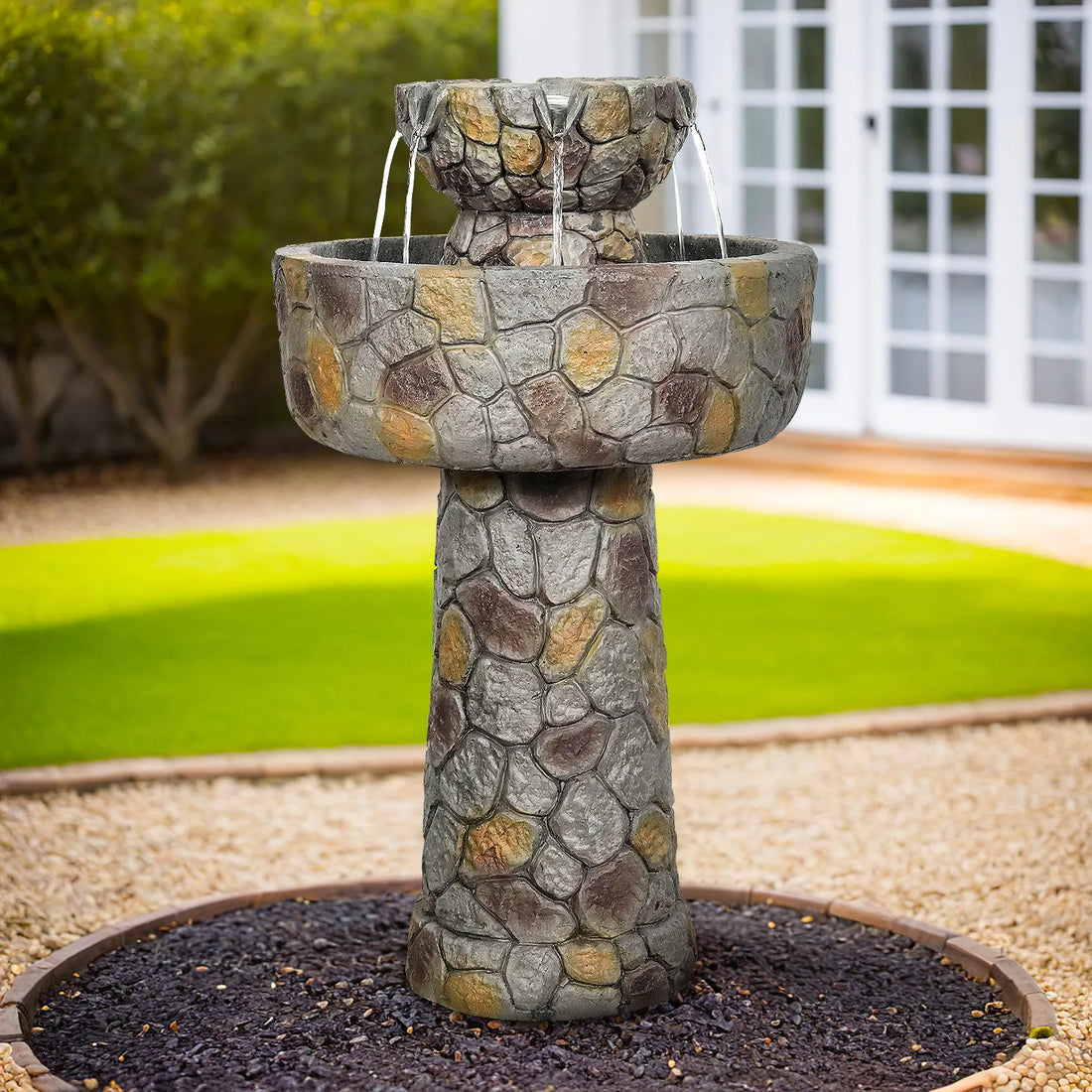 Decorative pedestal bird bath fountain with bubbling water flow on a dark mulch garden bed.
