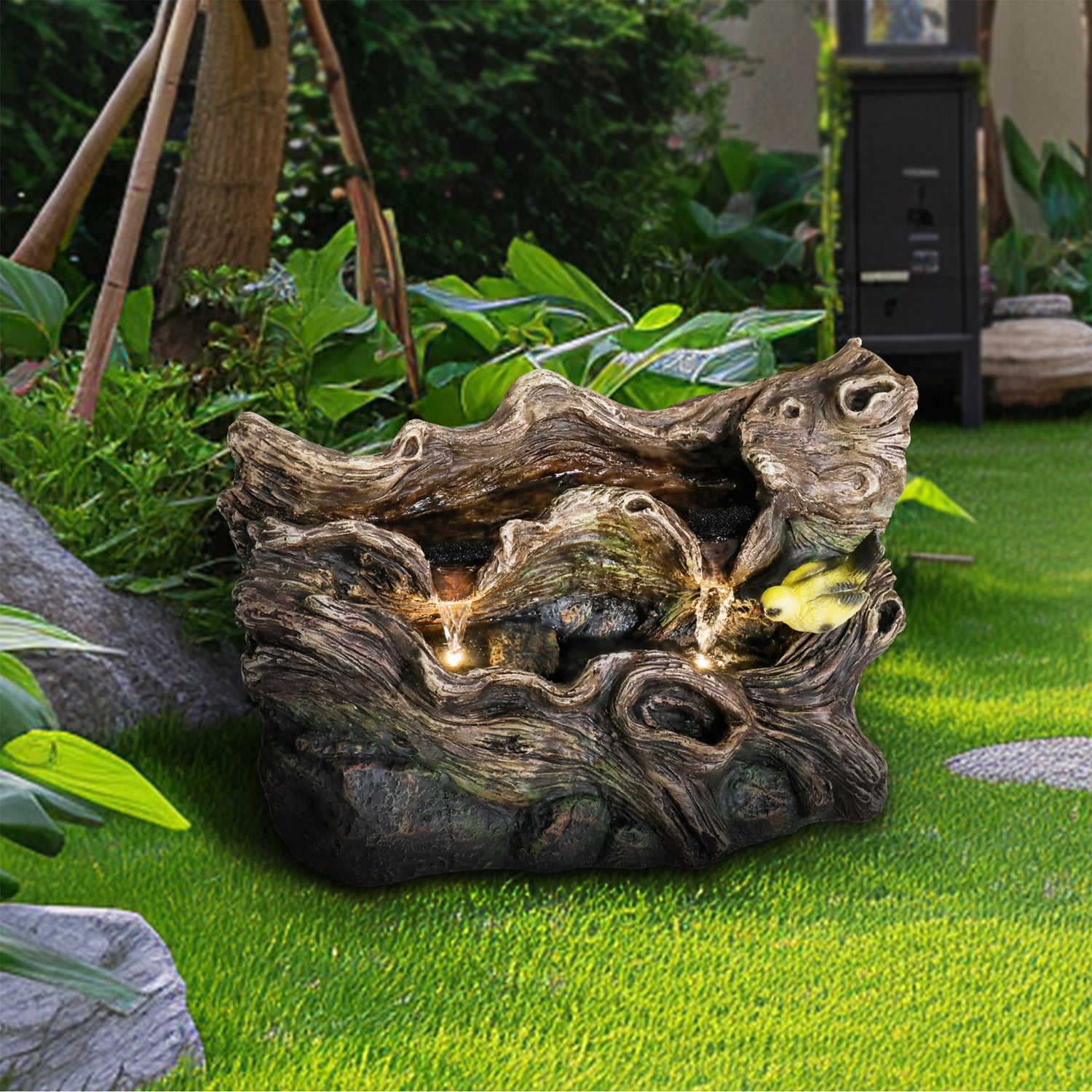 Realistic tree trunk and rock cascading fountain with LED lights and goldfinches on a garden lawn.
