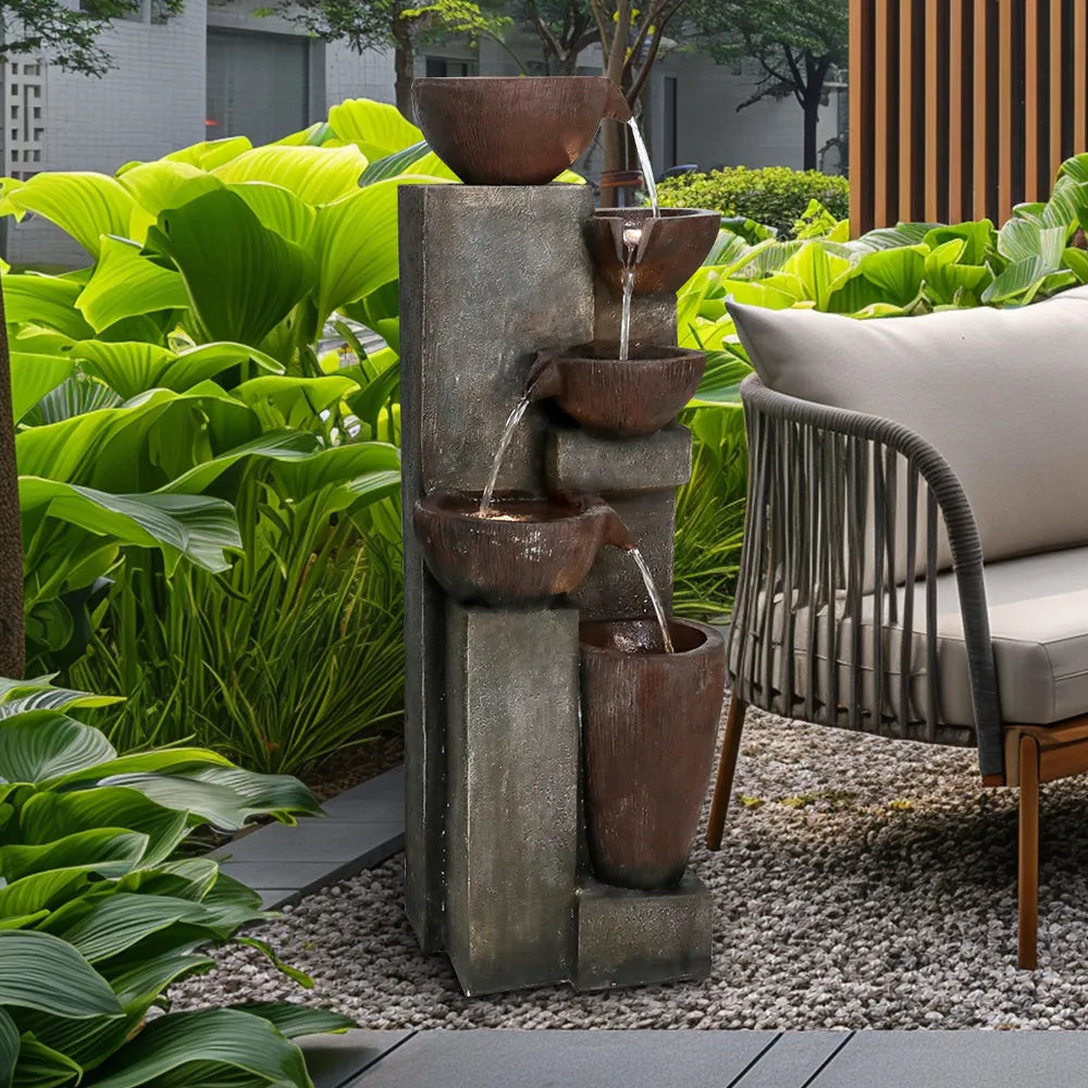 Simulated stone and copper tiered fountain viewed close-up, surrounded by lush green plants on a deck.