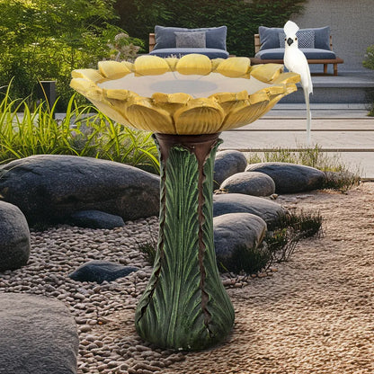 Sunflower-shaped birdbath with a yellow blossom basin and green leafy pedestal in a landscaped rock garden.