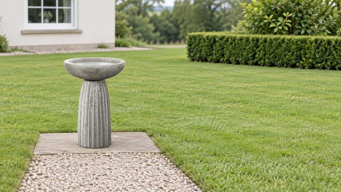 Fluted gray concrete pedestal bird bath on a flagstone pad beside a lawn, clipped hedge and house window behind