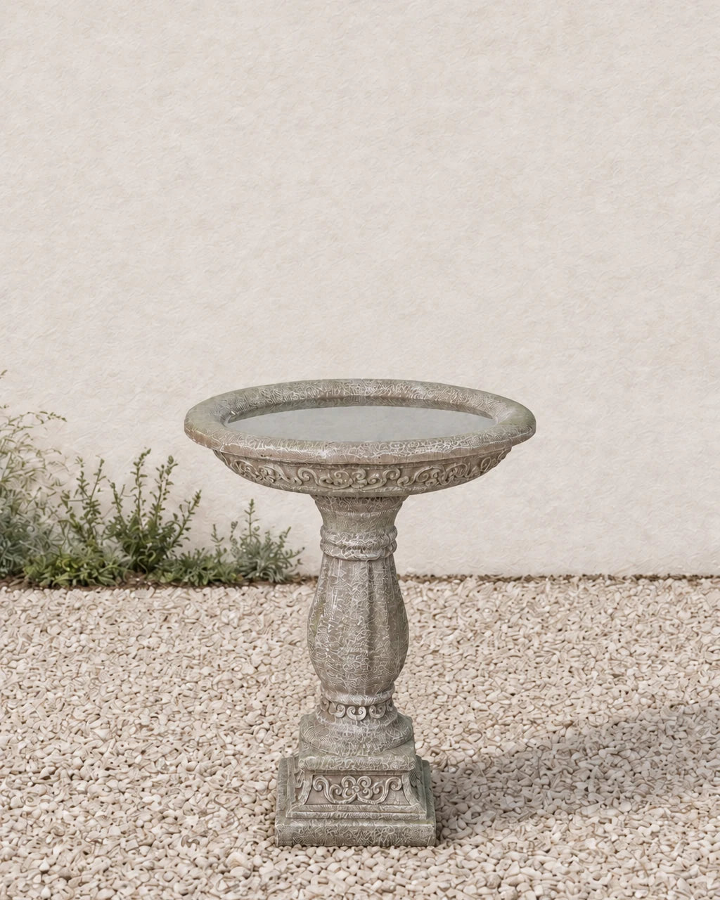 Antique ornate pedestal concrete bird bath on pale gravel in front of a planted bed