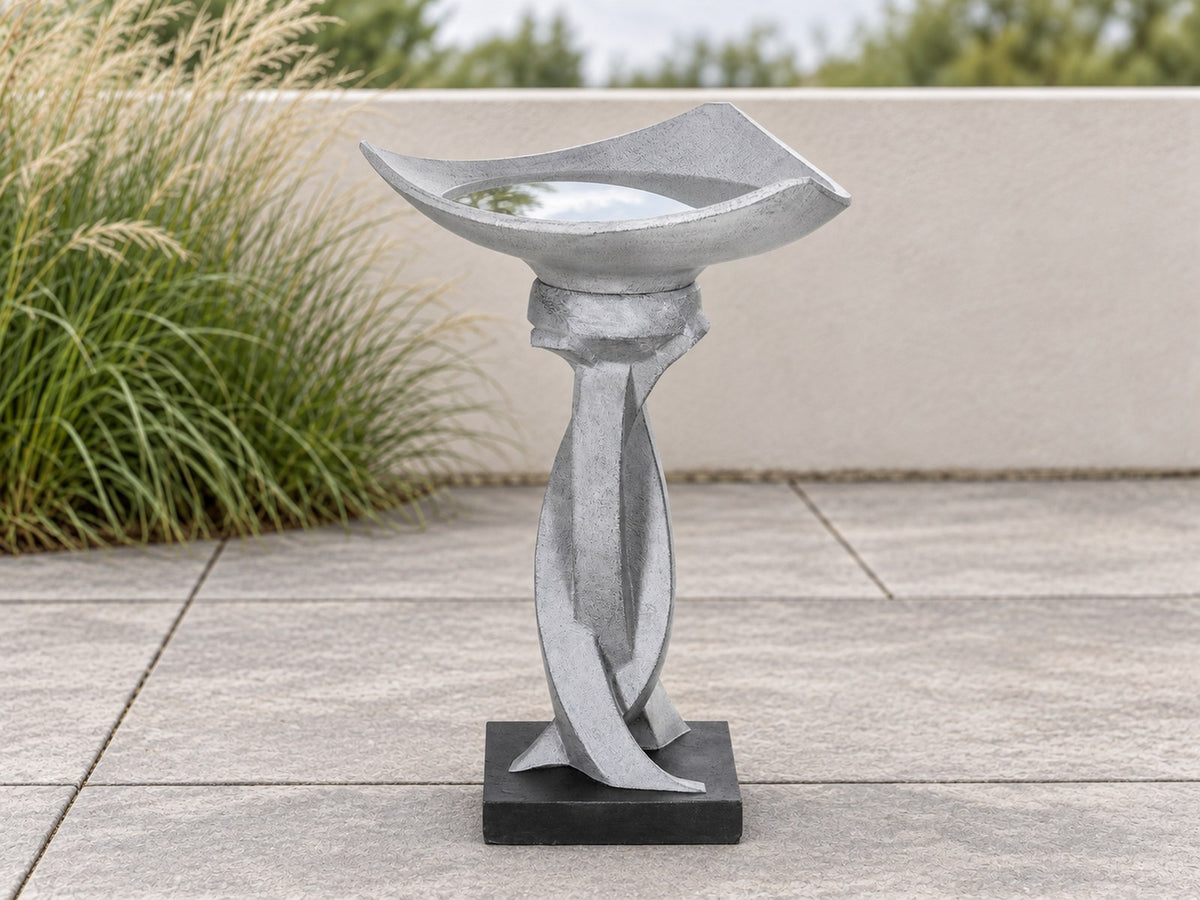 Sculptural twist concrete bird bath on a paved terrace with ornamental grasses behind