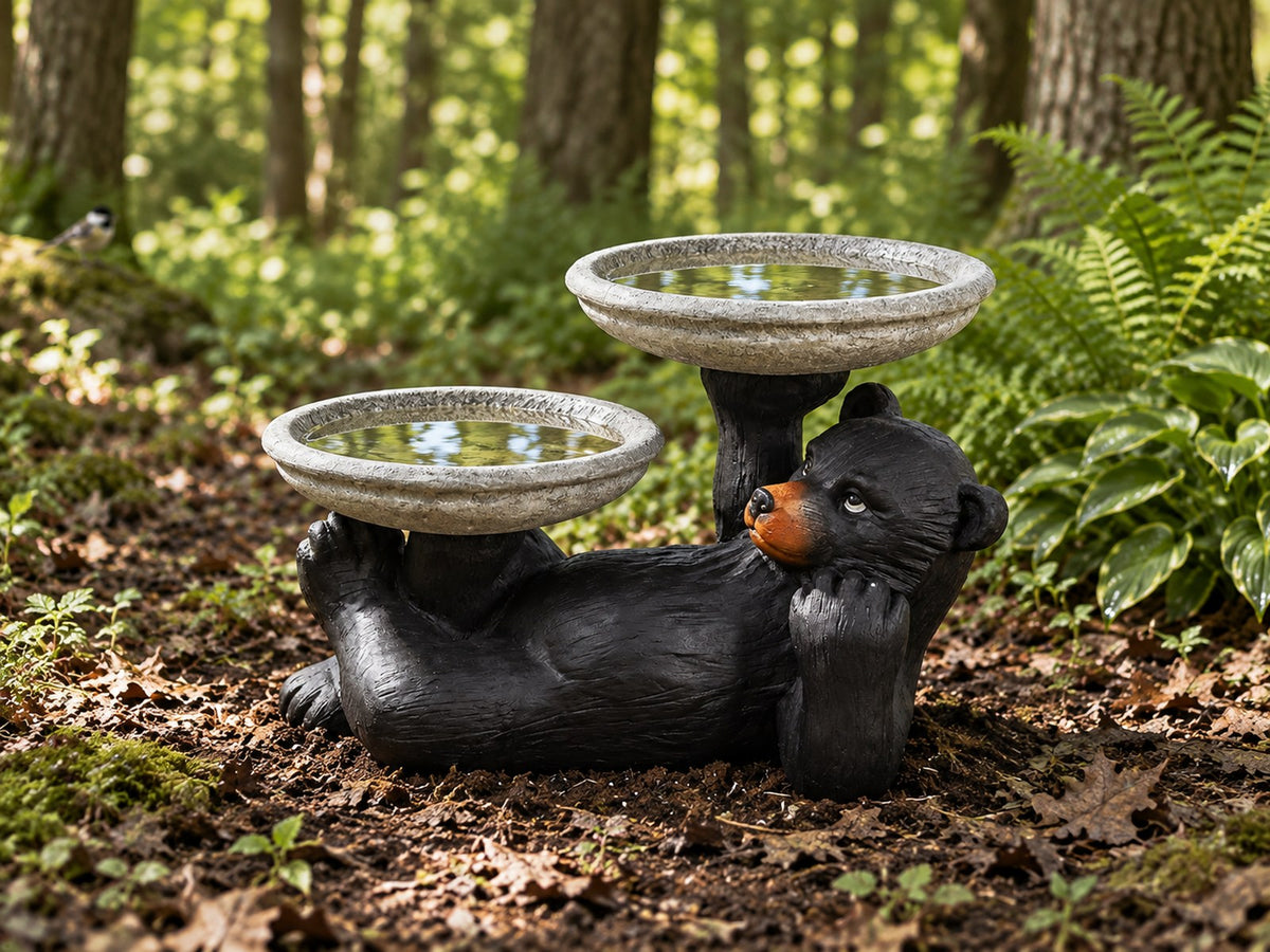 Black bear double bowl concrete bird bath on a woodland floor with ferns and dappled light