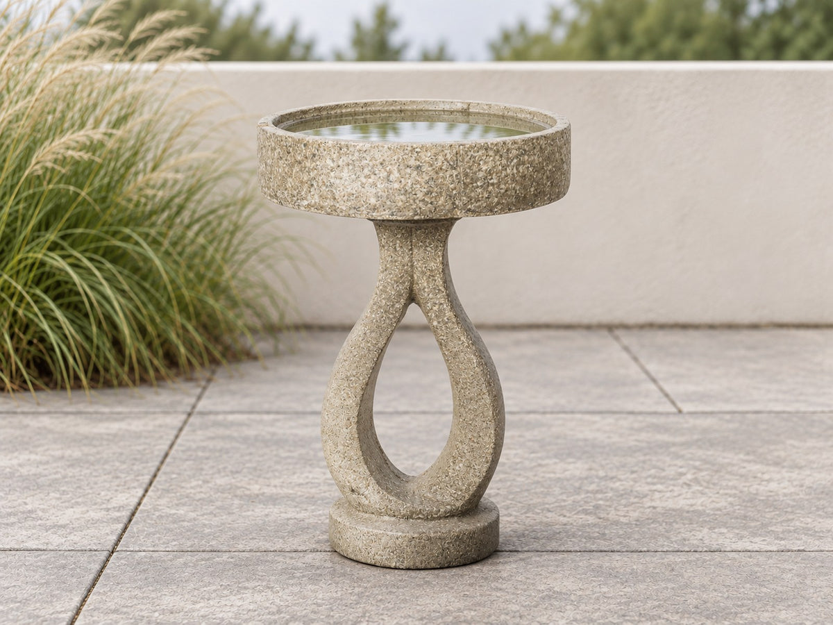 Open loop concrete bird bath on a paved terrace with ornamental grasses behind