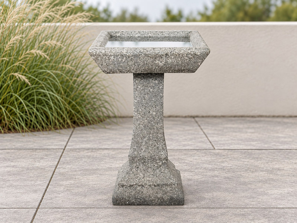 Square basin concrete bird bath on a paved terrace with ornamental grasses behind