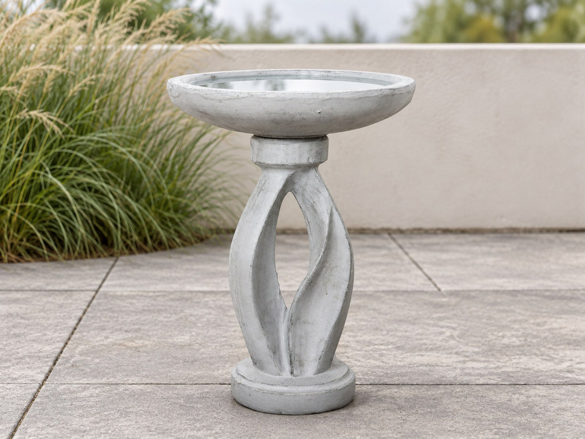 Twisted pedestal concrete bird bath on a paved terrace with ornamental grasses behind
