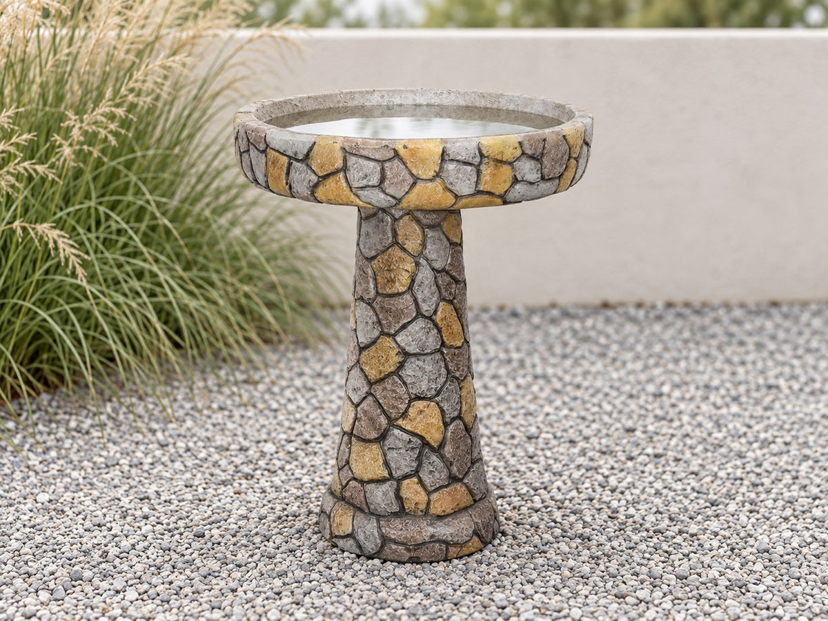 Mosaic stone concrete bird bath on fine gravel with ornamental grasses behind